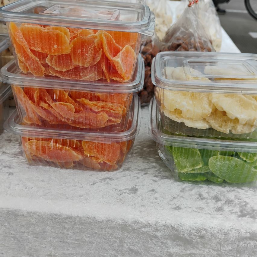 Drei Plastikbehälter mit orangefarbenen kandierten Früchten sind auf einem Tisch gestapelt, daneben befindet sich ein weiterer Behälter mit grünen kandierten Früchten.