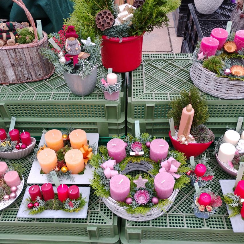 Various festive wreaths and candles, including a gnome figurine, displayed on green crates. Different colors and sizes of candles with floral decorations.