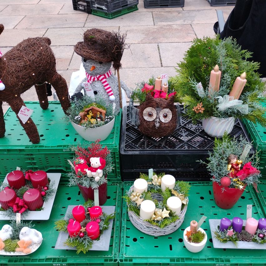 A display of festive decorations including wreaths, candles, and a snowman. The decorations are arranged on green crates and a black crate. The wreaths contain candles and are decorated with ribbons and flowers. The snowman has a price tag and a nativity scene.