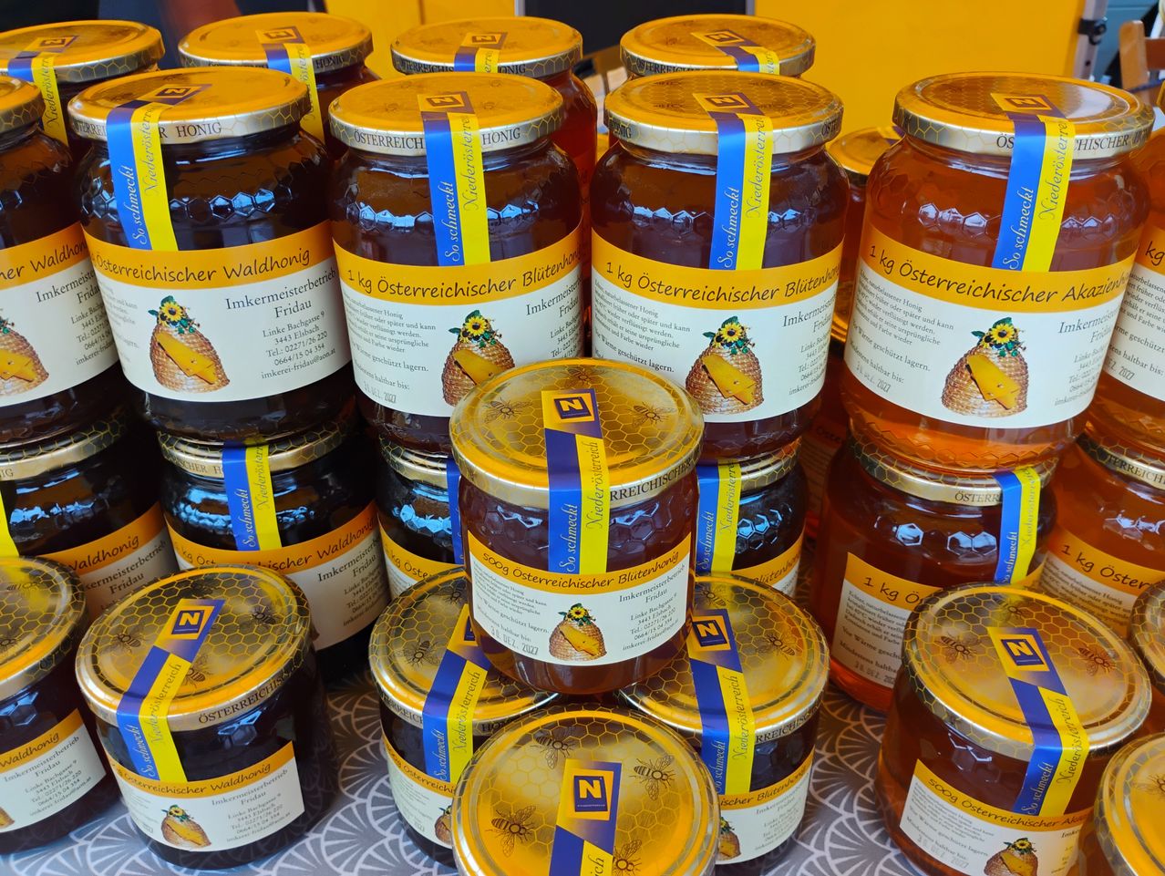 A collection of jars of honey with blue and yellow labels, stacked on a white surface. Each jar has a golden lid and contains honey with a honeycomb design.