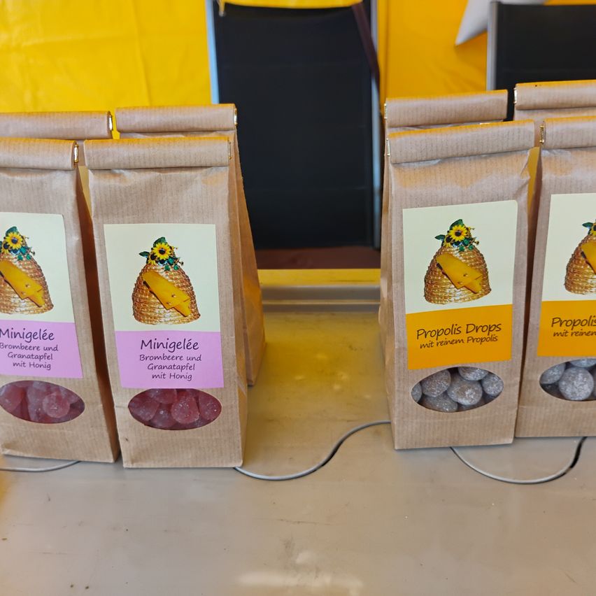 A row of brown paper bags with labels displaying different products. The first bag has a label that reads 'Minigelée' with a honeycomb and sunflowers. The second bag has a similar label but with 'Brombeer und Granatapfel mit Honig'. The third bag has a label that reads 'Propolis Drops mit reinem Propolis'. The fourth bag has a similar label but with 'Propolis mit reinem Propolis'.