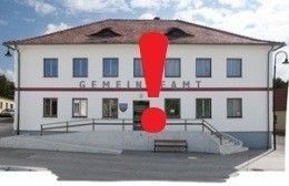Ein weißes Gebäude mit rotem Rand und einem roten Ausrufezeichen über dem Eingang heißt 'GEMEINDE'. Es gibt eine Bank und eine Treppe zum Eingang.