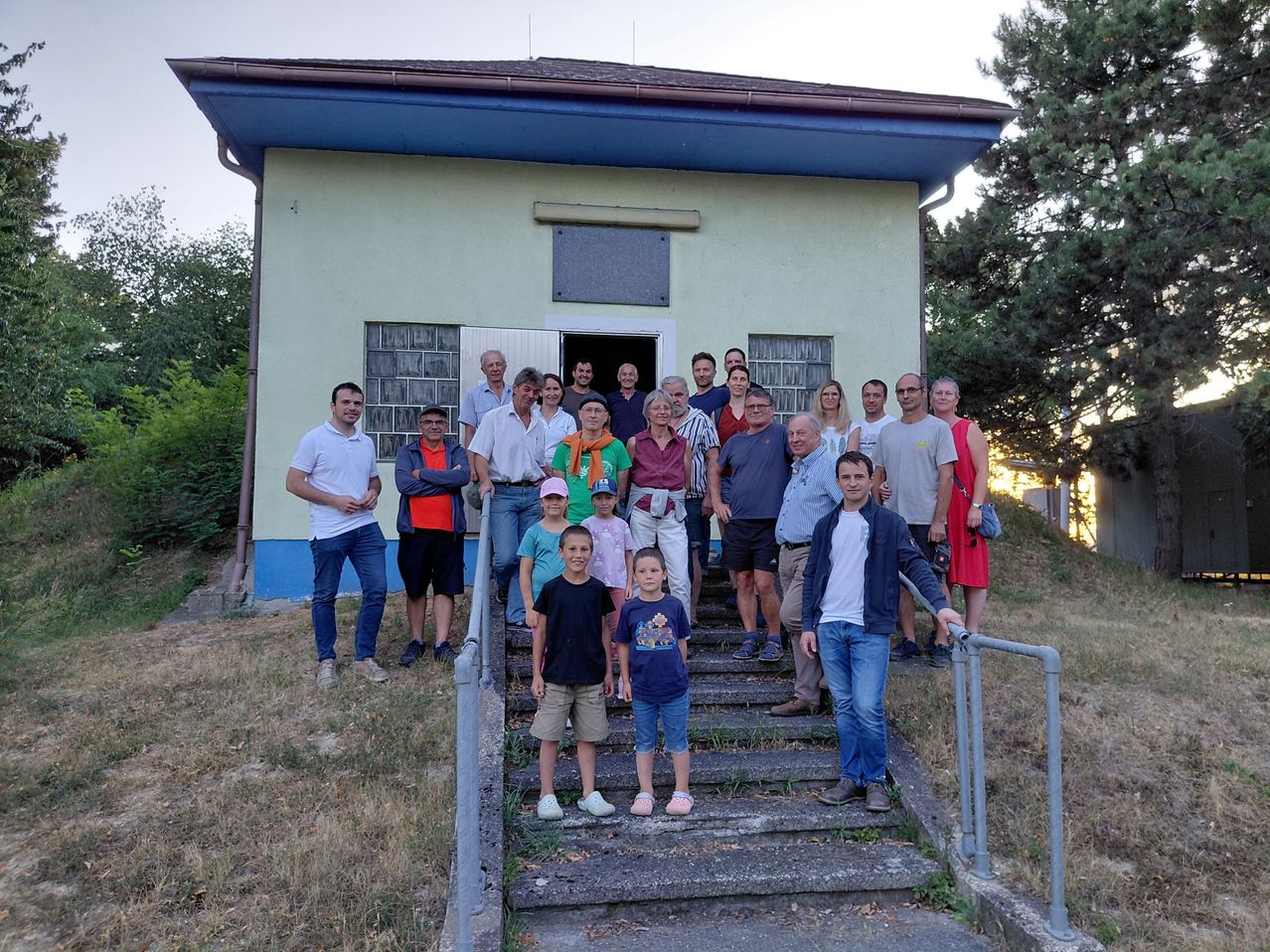 Eine Gruppe von Menschen, einschließlich Erwachsener und Kindern, steht auf den Stufen vor einem Gebäude mit blauem Dach und weißen Wänden.
