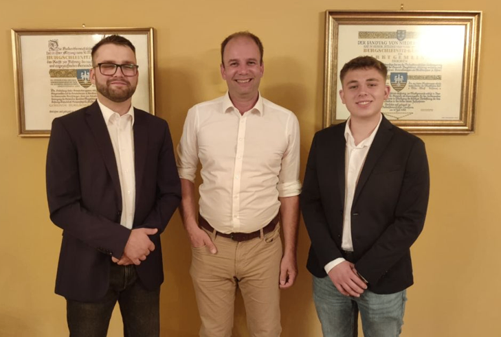 Drei Männer stehen zusammen vor einer gelben Wand mit gerahmten Zertifikaten. Der Mann auf der linken Seite trägt eine Brille und einen dunklen Anzug. Der Mann in der Mitte trägt ein weißes Hemd und Khakihosen. Der Mann auf der rechten Seite trägt einen dunklen Anzug und hat ein Lächeln im Gesicht.