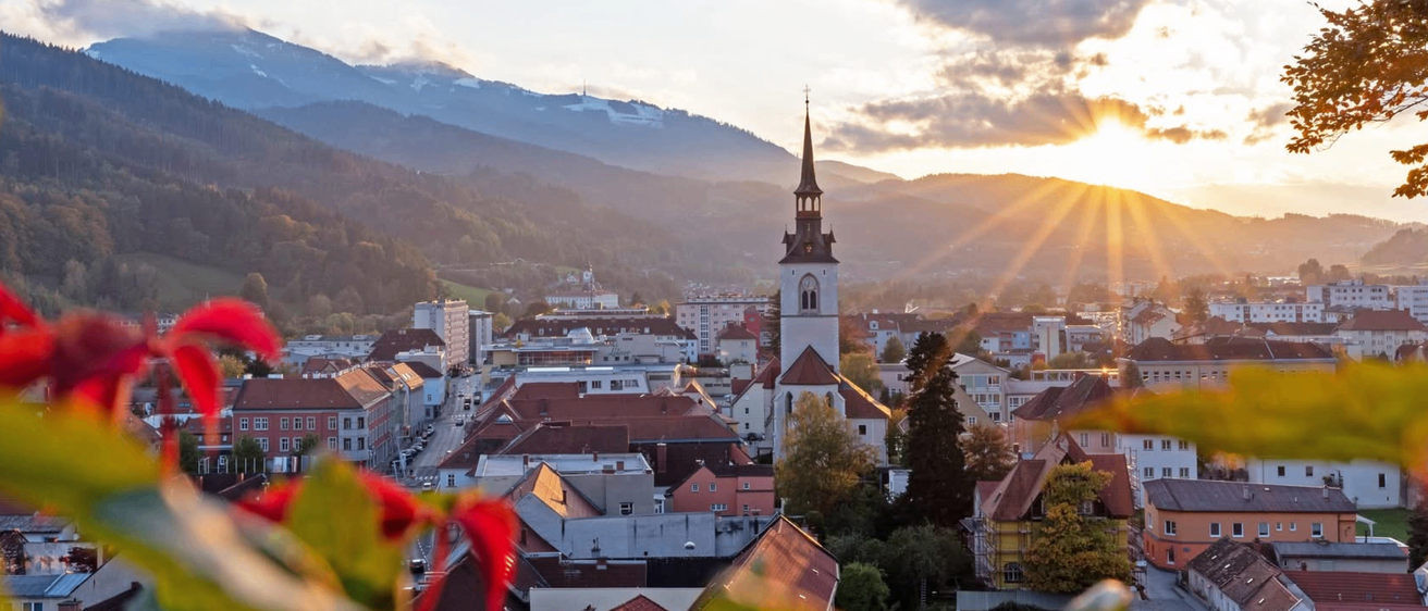 Luftaufnahme einer Stadt mit einem Kirchturm. Berge sind in der Ferne zu sehen. Die Sonne geht hinter den Wolken unter und wirft einen warmen Schein über die Landschaft.