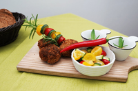 Ein Holzschneidebrett hält ein Gericht mit knusprigen Pastetchen und einem Gemüsespieß. Daneben stehen zwei Schüsseln mit Salat und einer roten Chilischote.