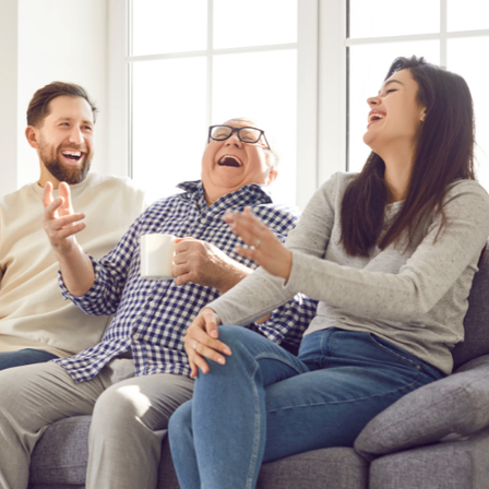 Drei Menschen sitzen auf einem Sofa. Ein Mann lacht und hält eine Tasse. Ein anderer Mann lächelt und gestikuliert mit der Hand. Die Frau lacht und schaut den Mann an.
