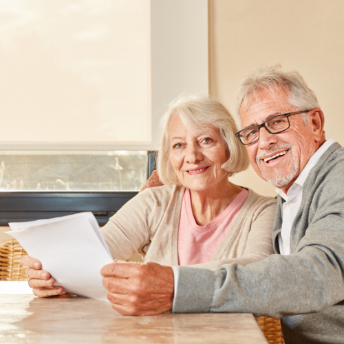 Ein älteres Paar sitzt an einem Tisch, lächelt und schaut auf ein Blatt Papier. Die Frau hält es, während der Mann sich vorbeugt. Sie scheinen zufrieden zu sein.