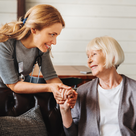 Eine Krankenschwester in einem grauen Dienstmantel hält die Hände einer älteren Frau mit kurzen weißen Haaren. Beide lächeln und schauen sich in einem Wohnzimmer an.