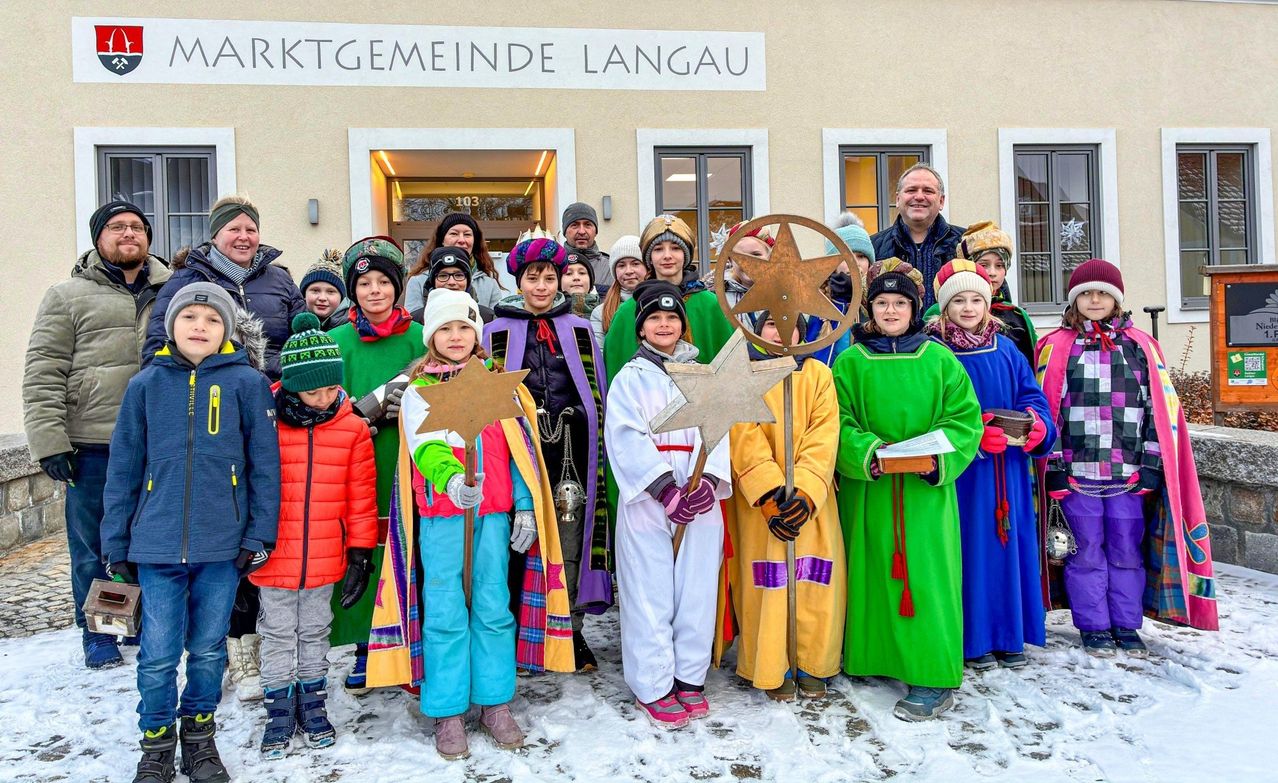 Eine Gruppe von Menschen in bunten Kostümen posiert für ein Foto vor einem Gebäude mit dem Text 'Krktgemeinde Langau'. Sie halten sternförmige Objekte in der Hand und tragen Winterkleidung.