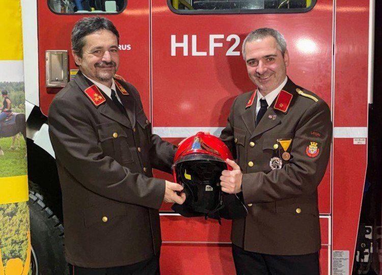 Zwei Männer in Uniform, die einen roten Helm halten, stehen vor einem roten Feuerwehrwagen mit der Aufschrift HLF2.