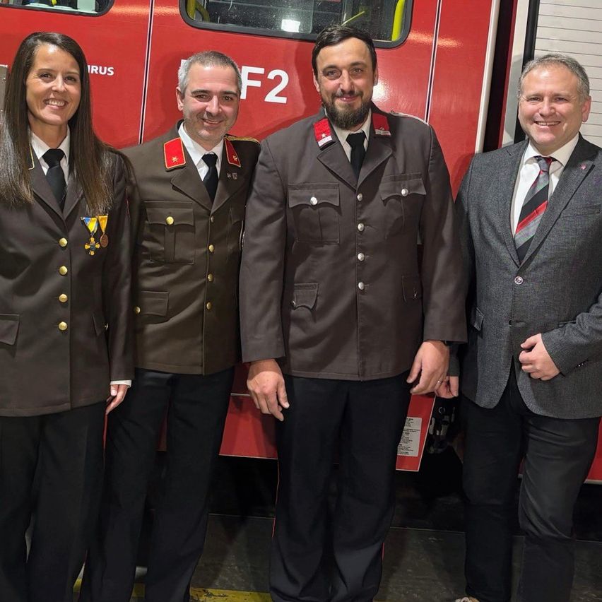 Vier Personen stehen vor einem roten Feuerwehrauto, tragen Uniformen und lächeln. Der Mann auf der rechten Seite hat eine gestreifte Krawatte.