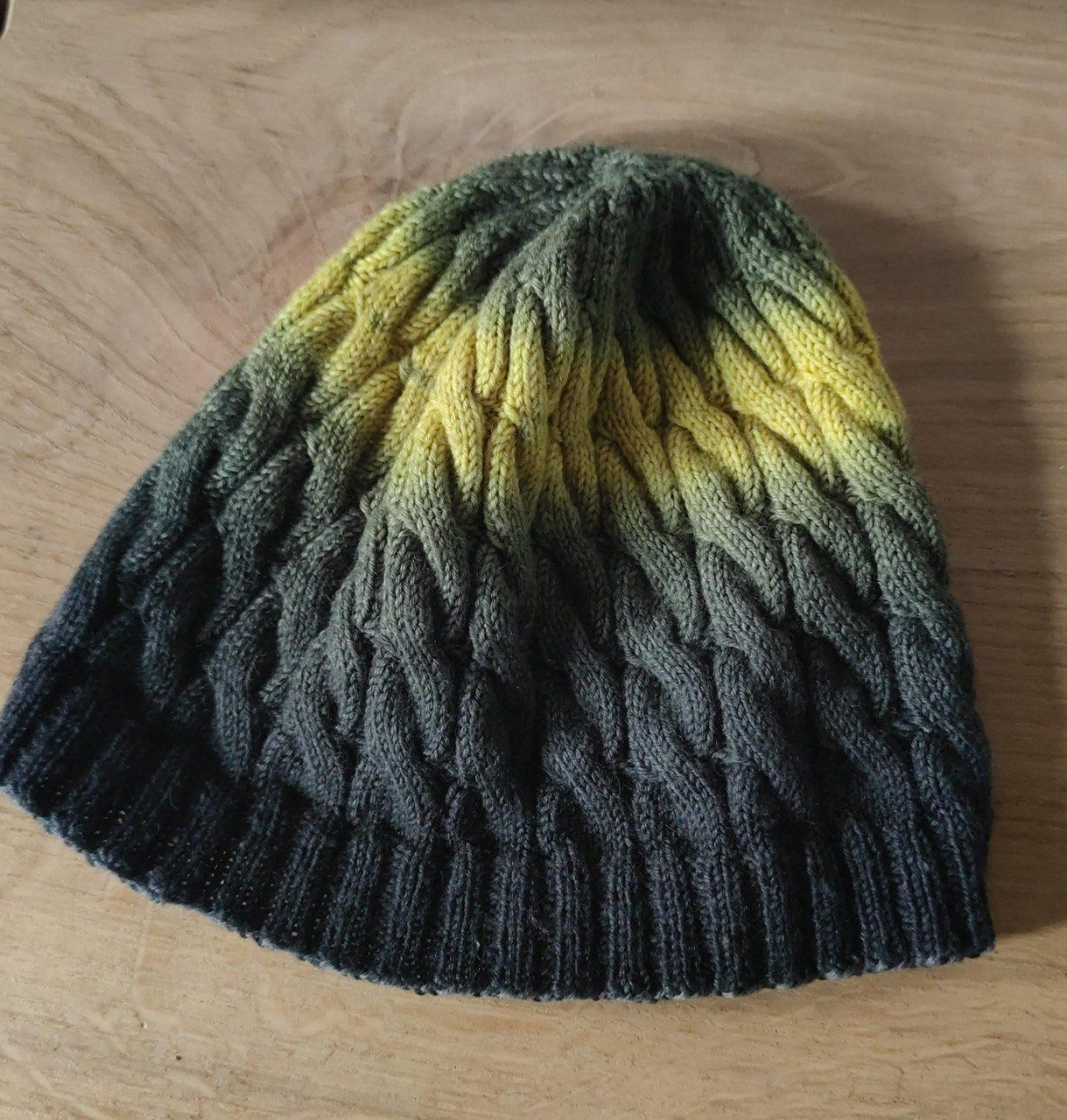Eine handgestrickte Mütze mit Farbverlauf von Gelb nach Schwarz wird auf einer Holzoberfläche präsentiert.
