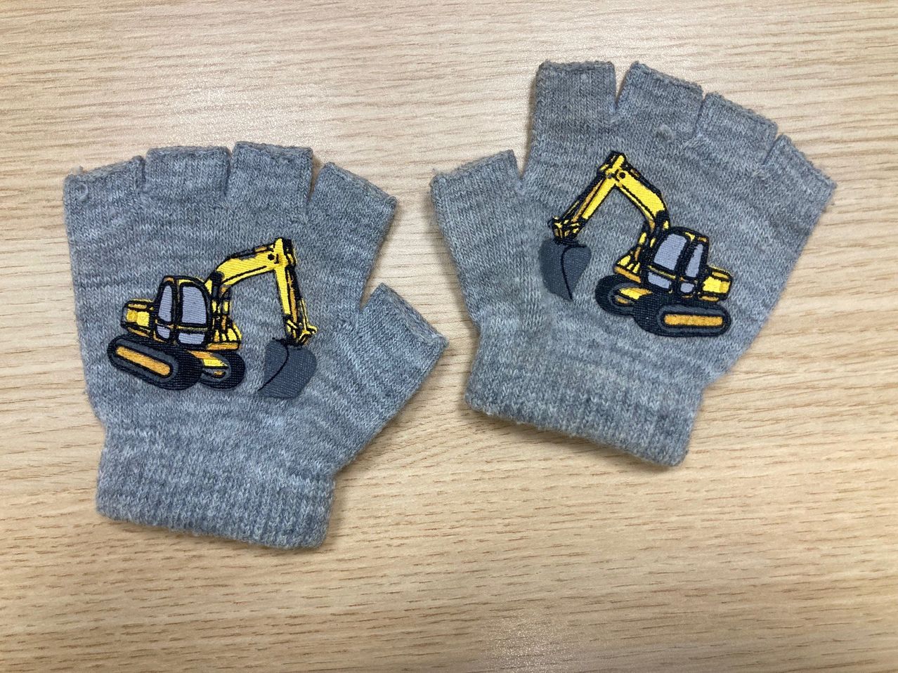 Zwei graue Handschuhe mit gelber Baggerstickerei auf einer Holzoberfläche.
