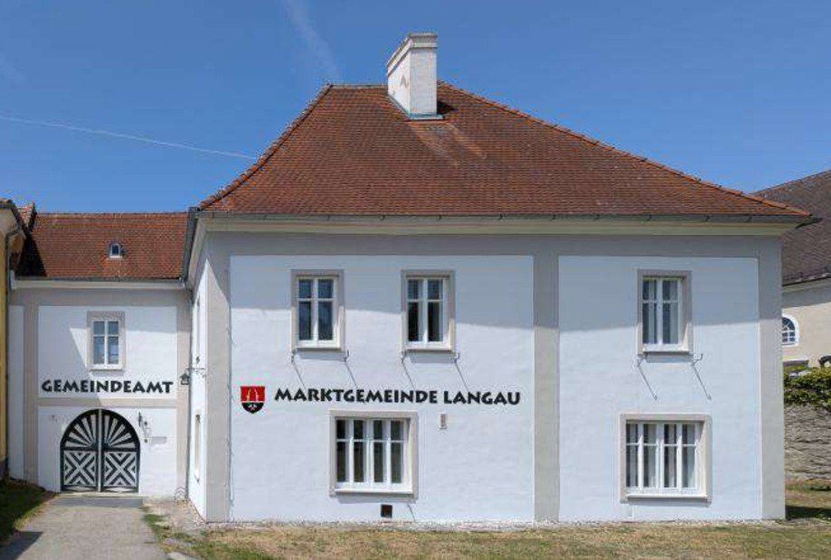 Ein weißes Gebäude mit einem braunen Dach und mehreren Fenstern, beschriftet mit 'Marktgemeinde Langau', steht unter einem klaren blauen Himmel.