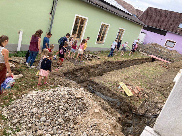 Mehrere Kinder und Erwachsene nehmen an einer Ausgrabung an einer Baustelle in der Nähe eines Hauses teil. Eine Frau hält eine Tasche.