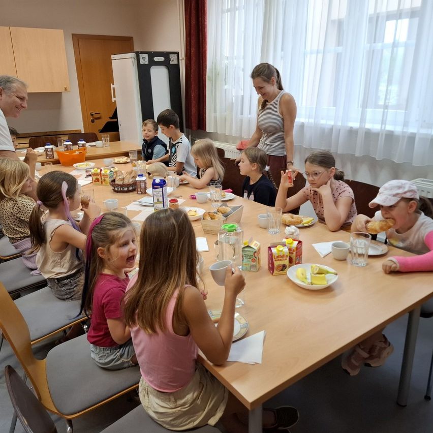 Eine Gruppe von Kindern sitzt um einen Tisch und isst Frühstück. Eine Frau steht hinter ihnen und serviert Essen. Der Tisch ist mit Tellern, Tassen und verschiedenen Lebensmitteln gedeckt.