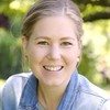 Eine lächelnde Frau mit blondem Haar, die eine Jeansjacke und eine Halskette trägt, posiert in einem Garten.