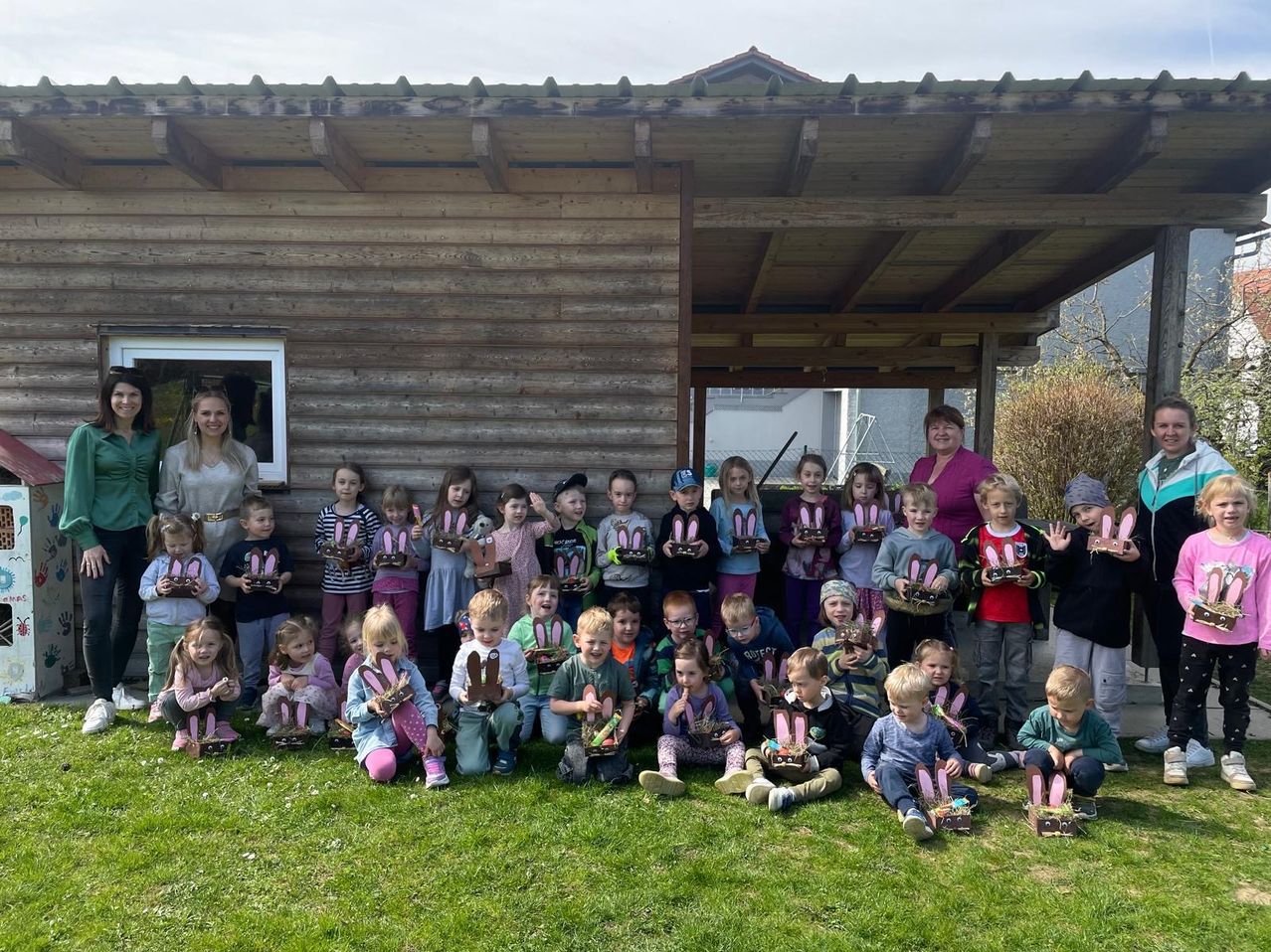Eine Gruppe von Kindern und Erwachsenen posiert mit Osterkörben auf einer Rasenfläche vor einem Holzgebäude.
