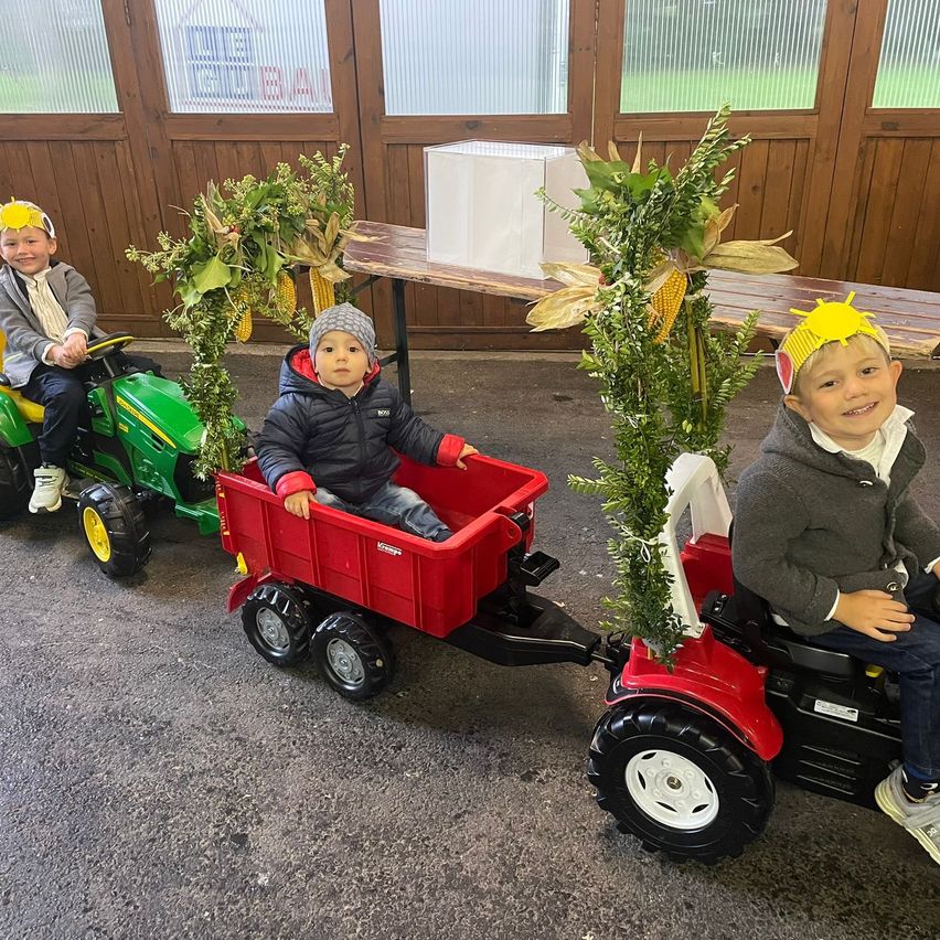 Drei kleine Kinder sitzen auf Spielzeugtraktoren mit Maisdekorationen, in einem Gebäude mit Glaswänden. Der Junge links trägt eine gelbe Krone.