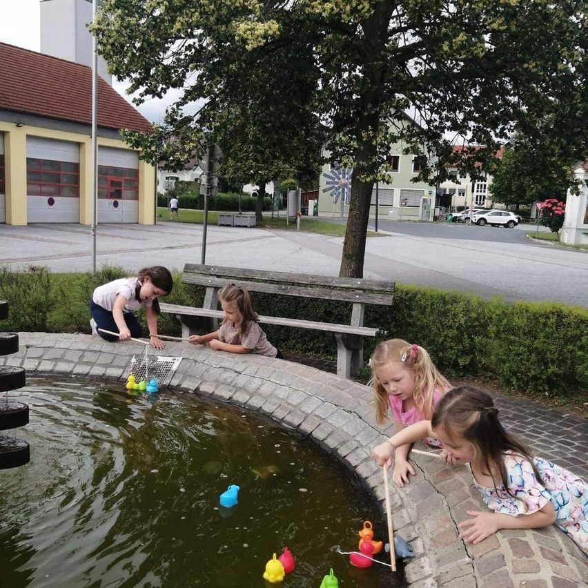 Vier Mädchen spielen mit bunten Entenspielzeug in einem Teich in der Nähe einer Bank und eines Gebäudes mit Garagentoren.