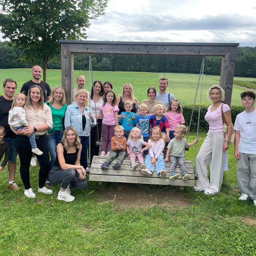 Eine große Familie ist für ein Foto auf einer Schaukel in einem Grasfeld versammelt, mit einem Feld und Bäumen im Hintergrund.