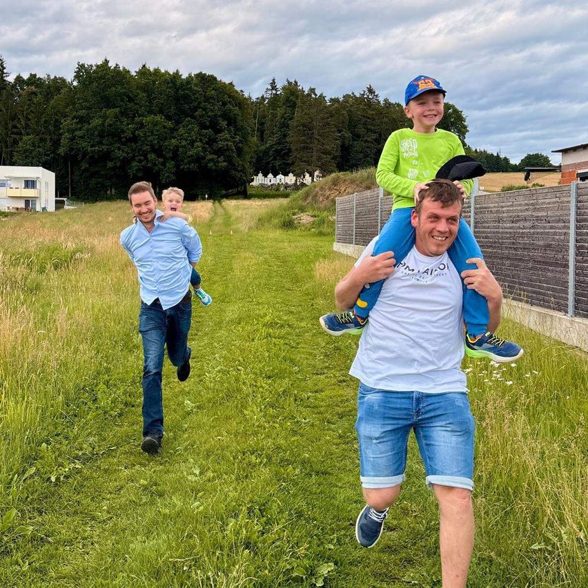 Drei Männer rennen auf einem Feld. Ein Mann trägt ein Kind auf seinen Schultern, während ein anderer ein Kind auf seinem Rücken trägt.