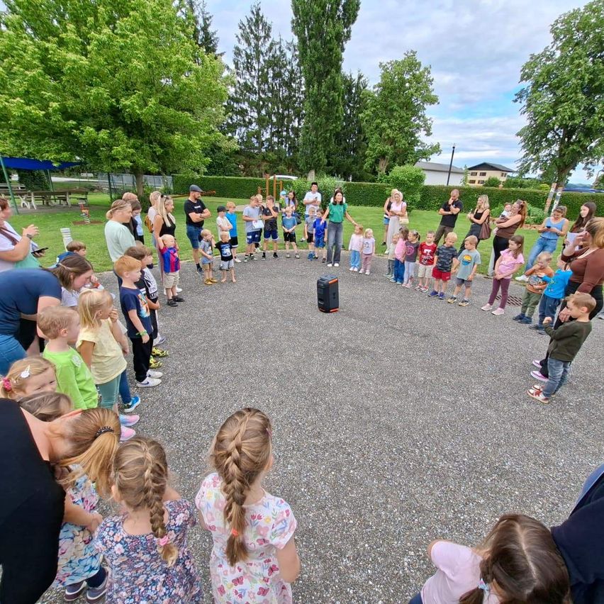 Eine Gruppe von Kindern und Erwachsenen ist in einem Kreis versammelt, wahrscheinlich für eine Schulveranstaltung. Sie stehen auf einem Kiesbereich mit einem kleinen schwarzen Lautsprecher in der Mitte. Bäume und Büsche befinden sich hinter ihnen, mit einem Gebäude in der Ferne.