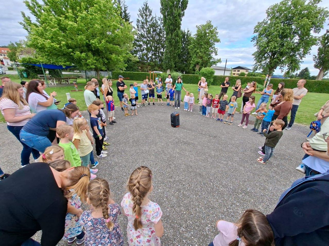 Eine Gruppe von Kindern und Erwachsenen ist in einem Kreis versammelt, wahrscheinlich für eine Schulveranstaltung. Sie stehen auf einem Kiesbereich mit einem kleinen schwarzen Lautsprecher in der Mitte. Bäume und Büsche befinden sich hinter ihnen, mit einem Gebäude in der Ferne.