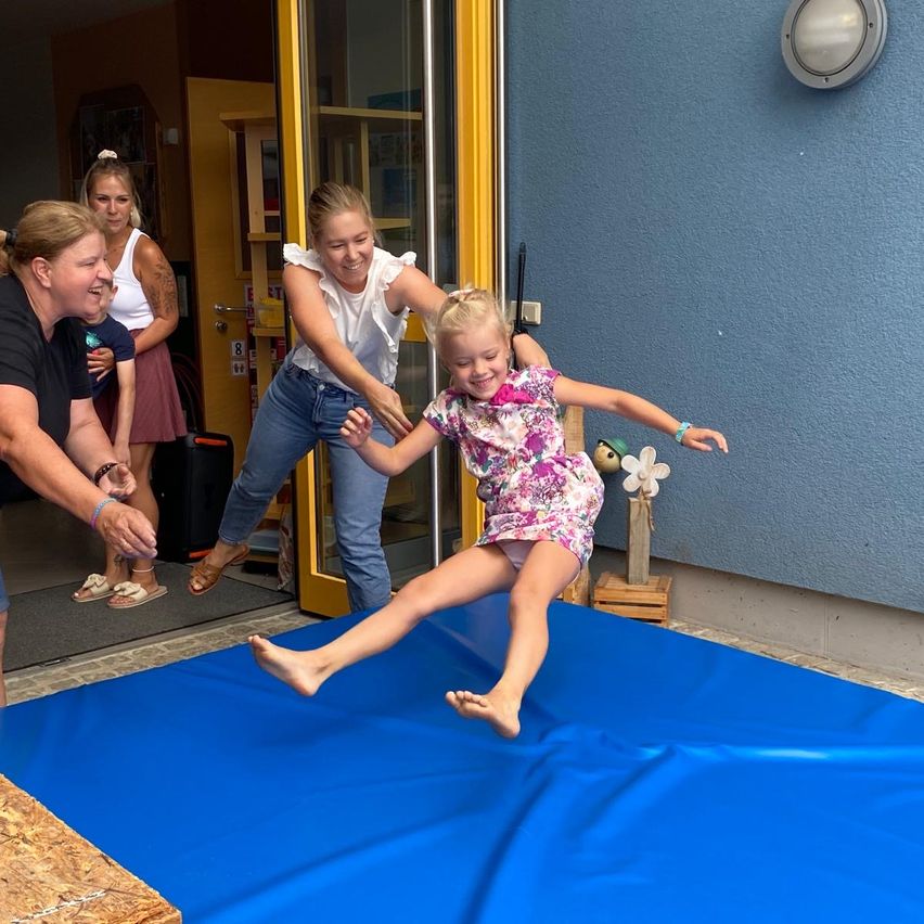 A young girl jumps on a blue mat while two women assist her, and others watch from the doorway.