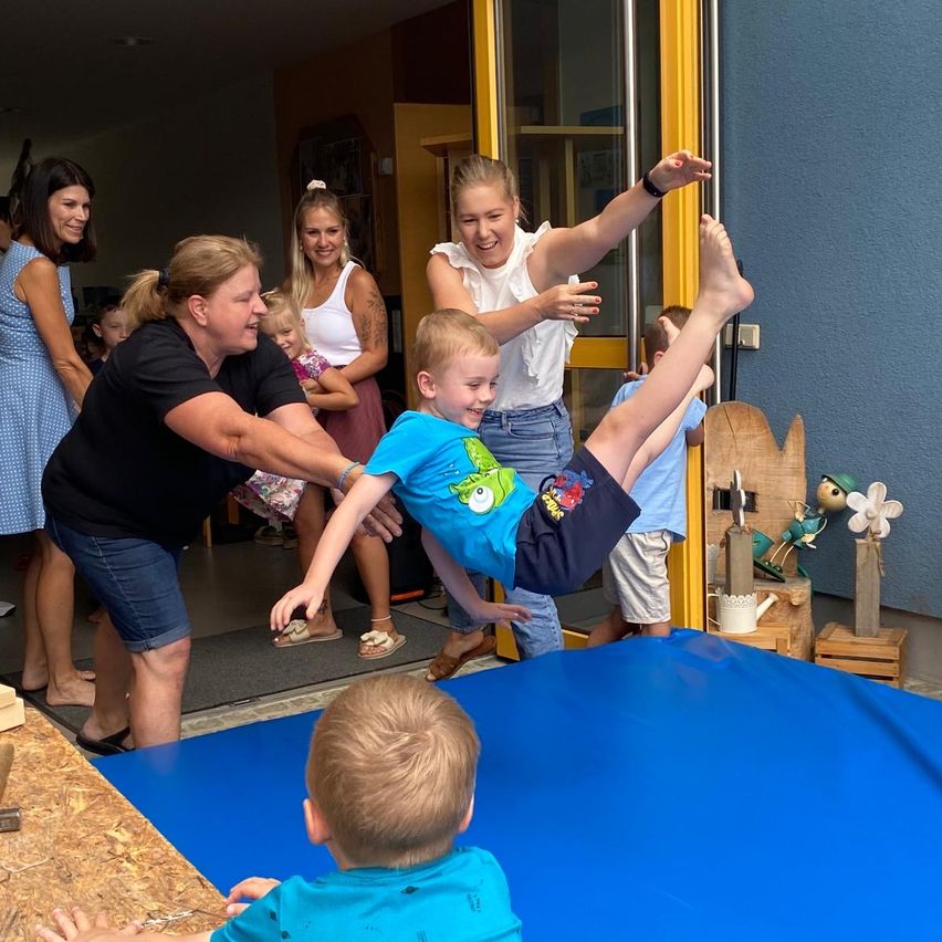 A group of children and adults are playing on a blue mat inside a room. A woman is helping a boy in a blue shirt to jump.