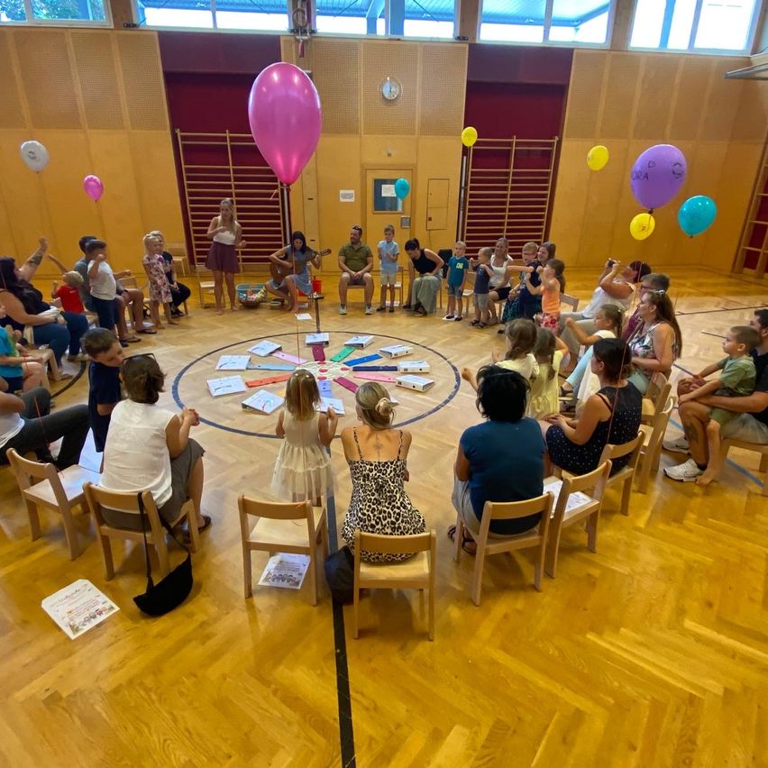 Eine Gruppe von Menschen, einschließlich Kindern, sitzen in einem Kreis auf Stühlen in einem Raum mit Holzböden und Ballons. Einige spielen Musikinstrumente.
