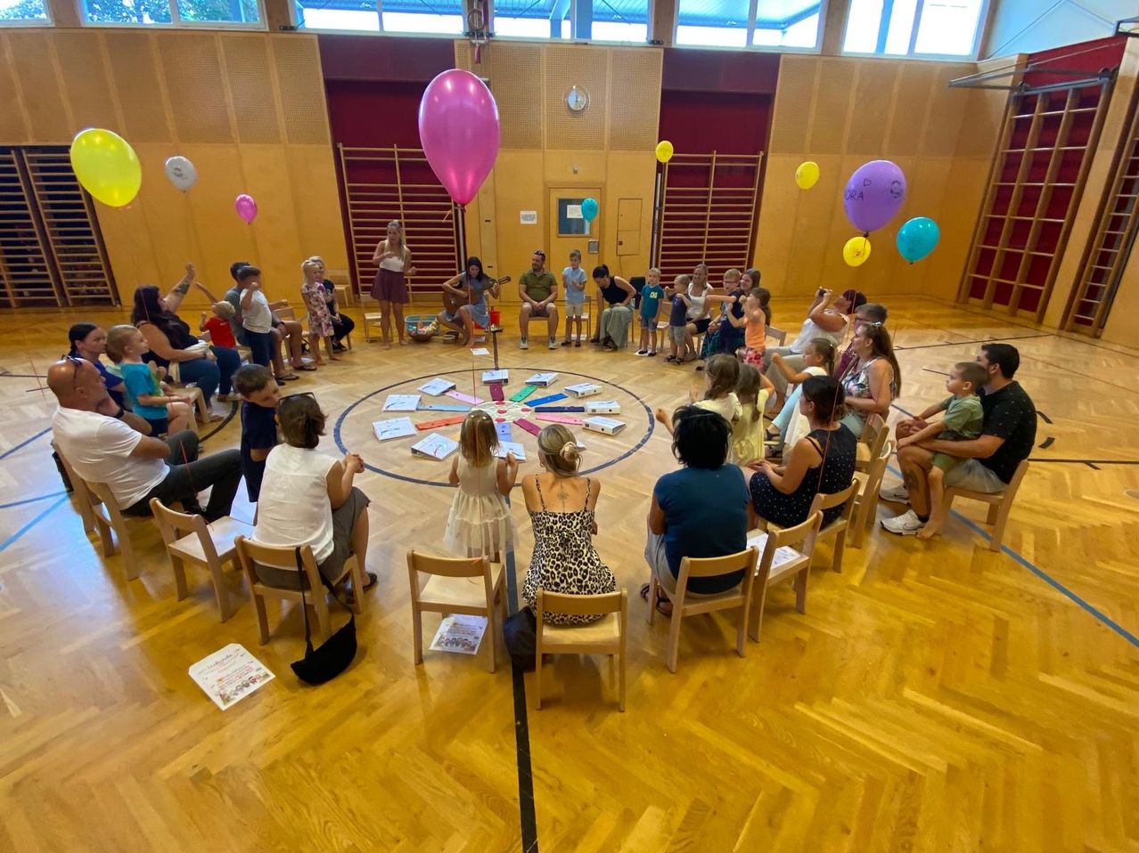 A group of people, including children, are sitting in a circle on chairs in a room with wooden floors and balloons. Some are playing musical instruments.
