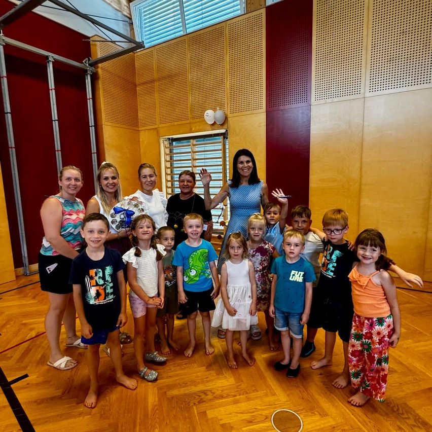 A group of children and adults are standing together in a room, smiling and posing for a photo. The children are wearing casual clothing, and the adults are standing behind them. The room has wooden floors and walls with a window.