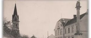 Schwarz-Weiß-Bild einer Dorfstraße mit einer Kirche, einem Denkmal und Leuten, die vor einem Gebäude stehen.