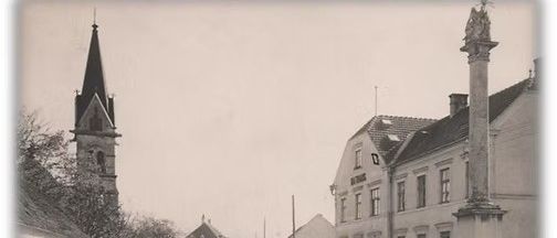 Schwarz-Weiß-Bild einer Dorfstraße mit einer Kirche, einem Denkmal und Leuten, die vor einem Gebäude stehen.