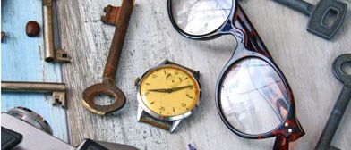 Eine Sammlung alter Gegenstände, darunter Schlüssel, eine Uhr, eine Brille, eine Kamera, eine Schachtel und Blumen auf einem Holztisch.