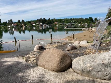 Ein Seeufer mit einem Wasserbrunnen, umgeben von Felsen und Bäumen. Ein Boot ist auf dem Wasser zu sehen. In der Ferne stehen Häuser in Reihe nahe den Bäumen.