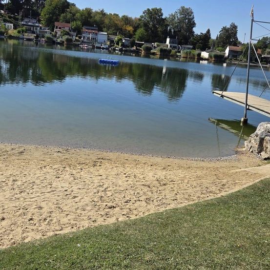 Ein ruhiger See mit einem sandigen Ufer und einem Steg auf der rechten Seite, umgeben von Bäumen und Häusern im Hintergrund.
