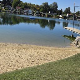 Ein ruhiger See mit einem sandigen Ufer und einem Steg auf der rechten Seite, umgeben von Bäumen und Häusern im Hintergrund.