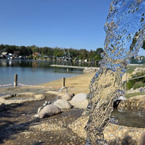 Eine Nahaufnahme eines Wasserbrunnens, der Wasser in die Luft sprüht, mit einem See und einem Strand im Hintergrund.