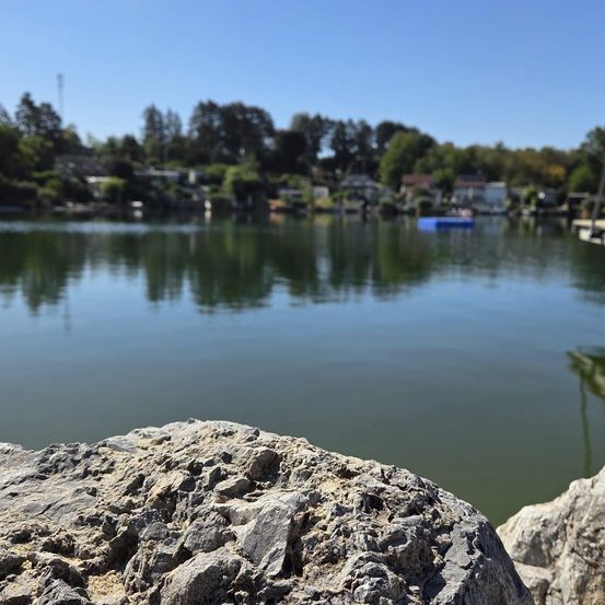 Ein ruhiger See mit einem felsigen Ufer und einem blauen aufblasbaren Boot auf dem Wasser. Bäume und Häuser sind im Hintergrund zu sehen.