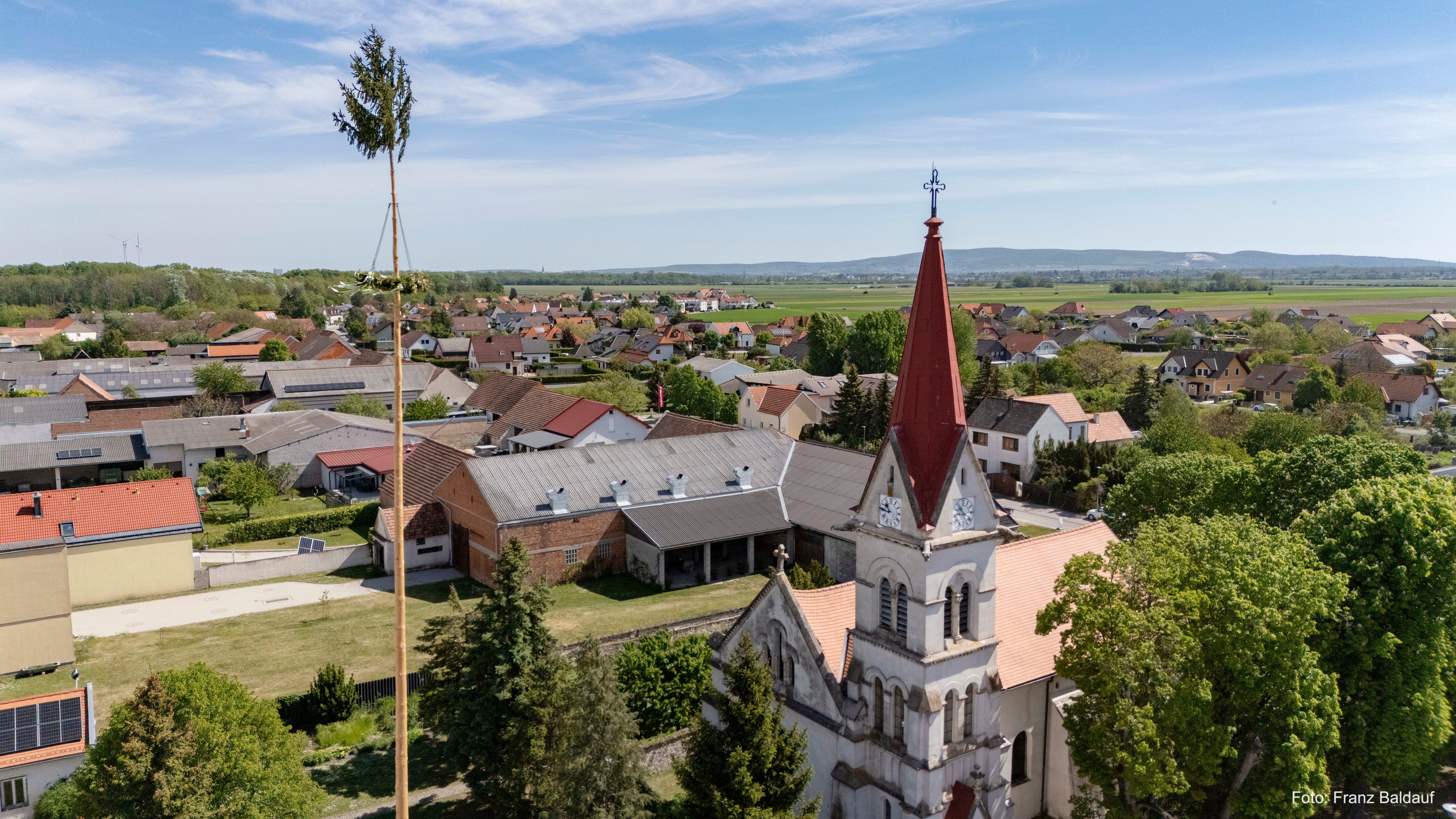 Luftaufnahme eines Dorfes mit einer Kirche, rotem Kirchturm und Häusern. Ein hoher Baum steht links, und ein grünes Feld ist in der Ferne.
