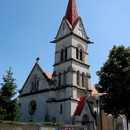 Eine gotische Kirche mit einem roten Turm und einer Uhr auf jeder Seite, umgeben von Bäumen und einer Straßenlampe.