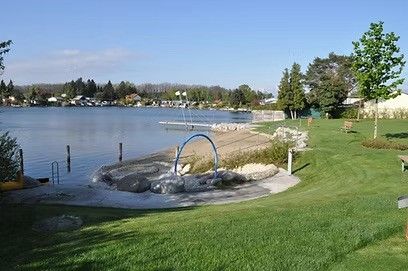 Eine ruhige Uferzone am See mit einem Sandstrand, einem Spielplatz und einem Grasfeld. Bäume und eine Bank sind zu sehen, mit einem Bootsanleger in der Ferne. Der Himmel ist klar.