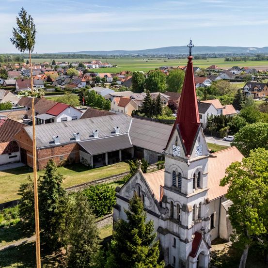 Ein Luftbild einer Stadt mit Häusern, einer Kirche und einem hohen Baum. Die Kirche hat einen roten Turm und eine Uhr. Bäume und Pflanzen umgeben das Gebiet.