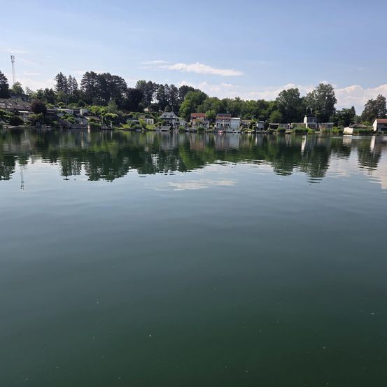 Ein ruhiger See spiegelt den Himmel und die Bäume wider. Am Ufer sind Häuser und Gebäude zu sehen, umgeben von Grün.