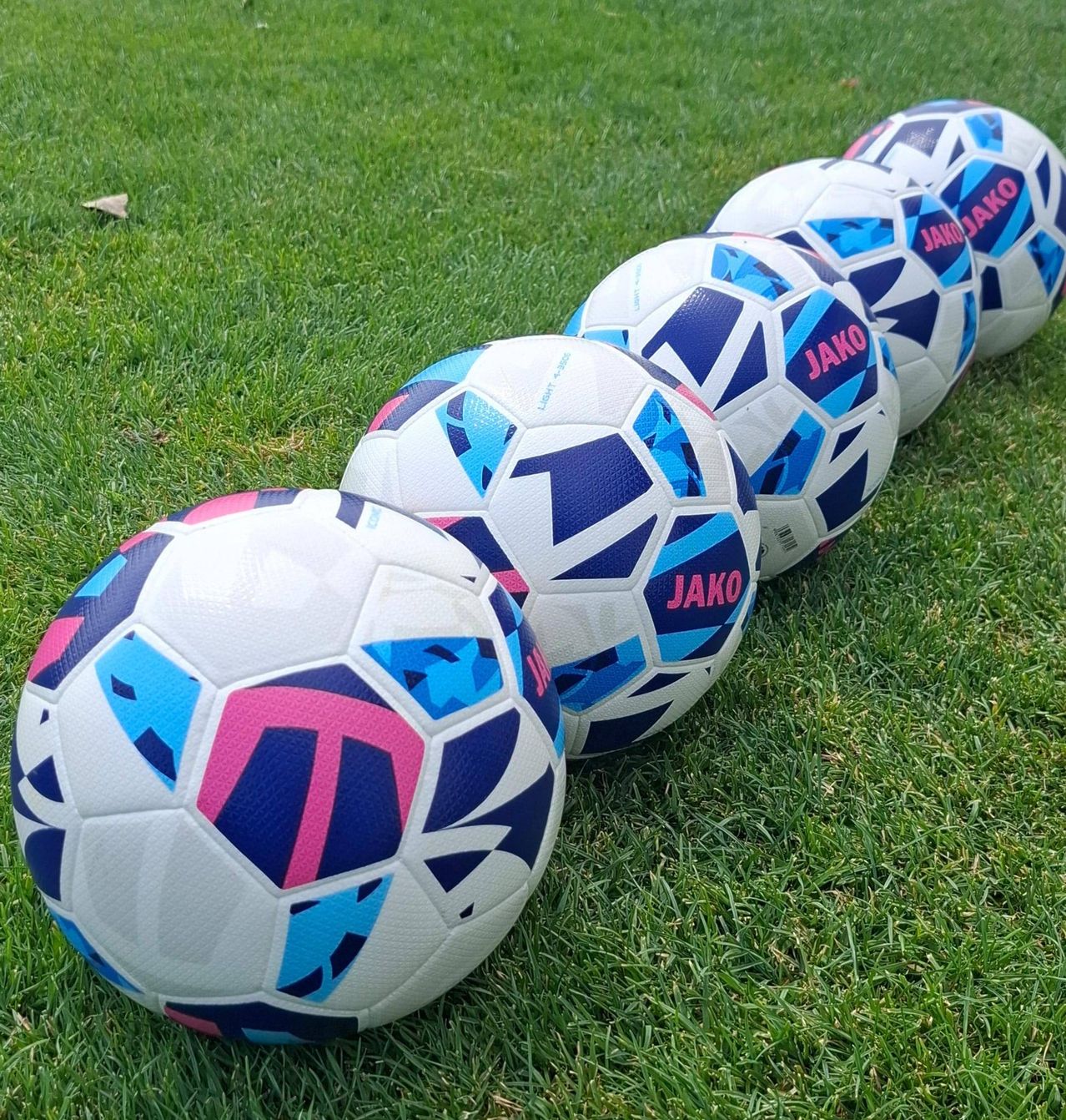 Fünf Fußballbälle mit blauen, rosa und weißen Designs sind auf einem Grasfeld aufgereiht. Jeder Ball hat den Namen Jako darauf gedruckt.
