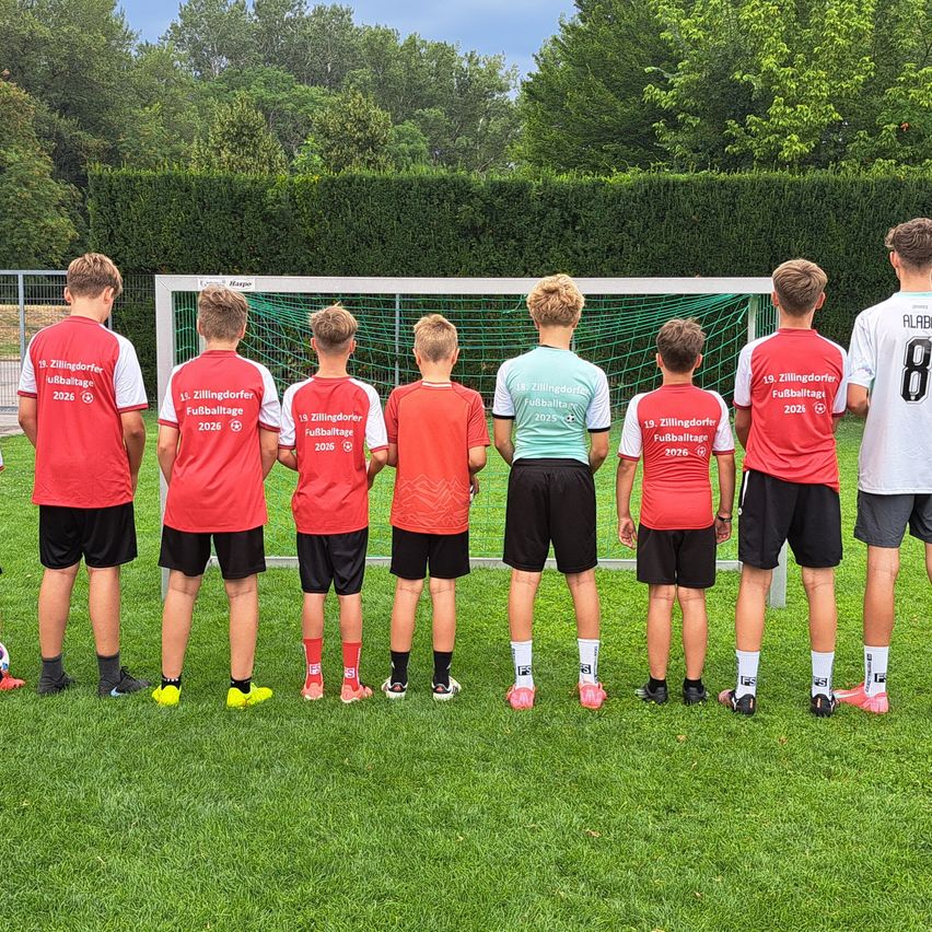 Eine Gruppe junger Fußballspieler steht in einer Reihe vor einem Tor, trägt rote und weiße Trikots mit Nummern und Namen. Der Rasen ist grün mit einer Hecke im Hintergrund.