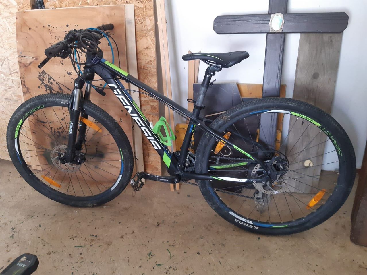 Ein schwarz-grünes Mountainbike steht in einer Garage mit Holzwänden. Das Fahrrad hat einen schwarzen Sattel und einen Flaschenhalter am Rahmen.